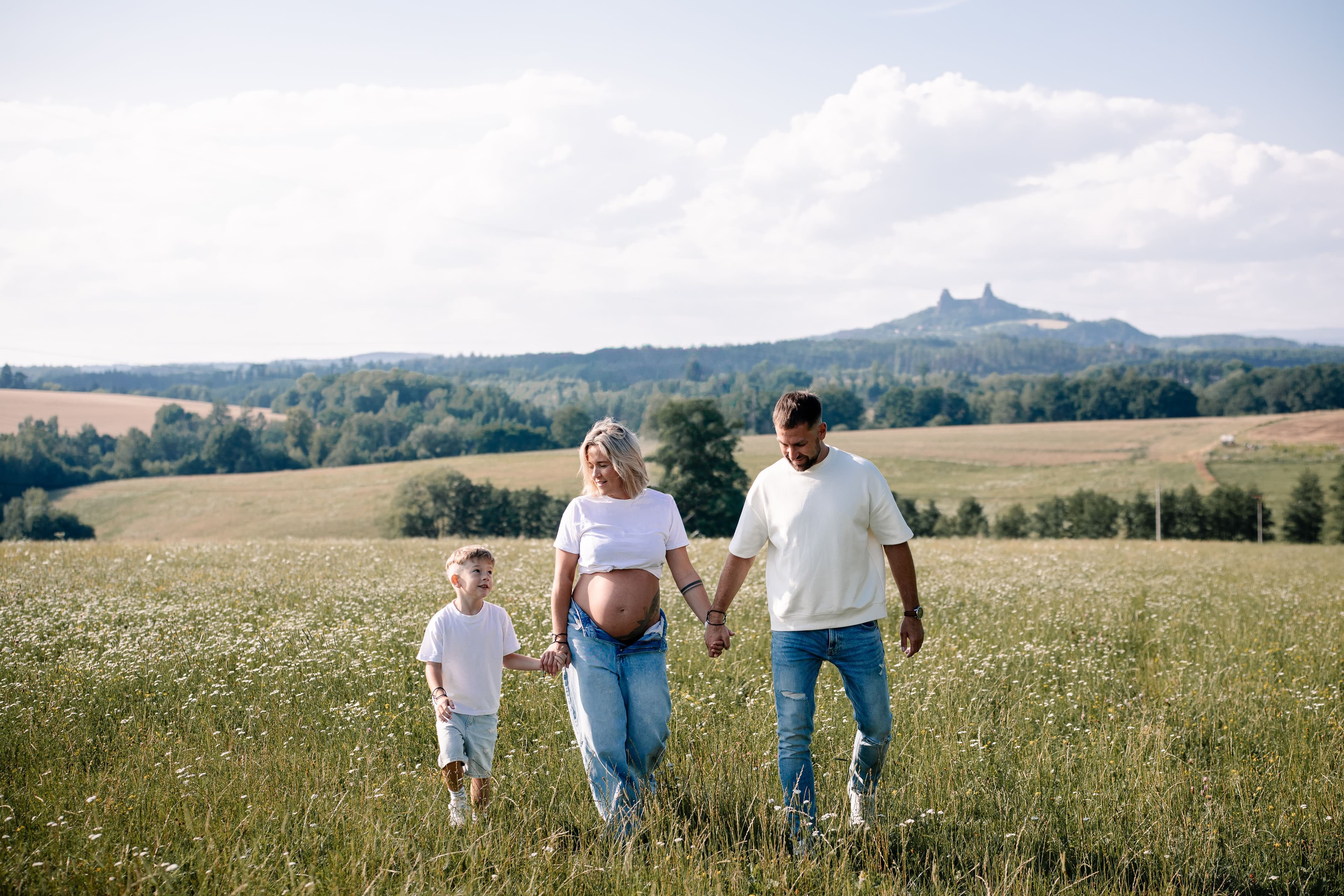Rodinné focení, těhotná maminka, tatínek a dítě se drží za ruce a jdou po rozkvetlé louce s výhledem na hrad Trosky, Michaela Čížková, fotografka, Mladějov, Český ráj